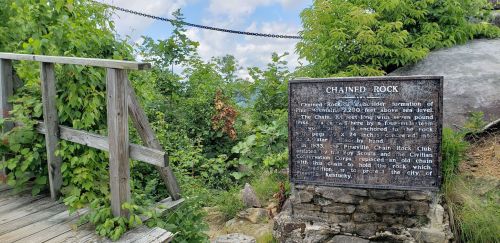 Sign about Chained Rock, a geological formation, with surrounding greenery and a wooden walkway in the background.