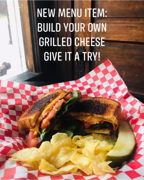 A grilled cheese sandwich with lettuce and tomato, served with chips and a pickle in a red checkered basket.