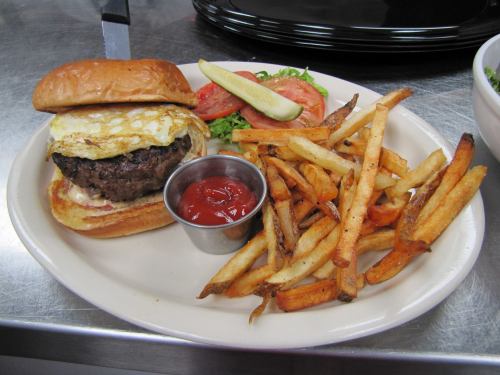 A juicy burger with a fried egg on a bun, served with fries, ketchup, lettuce, tomato, and a pickle on the side.
