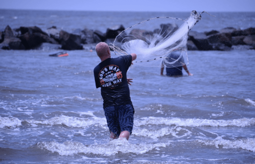 A person casts a fishing net into the ocean while wading through the water, with rocky shore in the background.