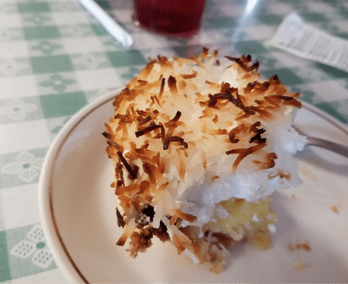 A slice of coconut cream dessert with a toasted coconut topping on a white plate, set on a checkered tablecloth.