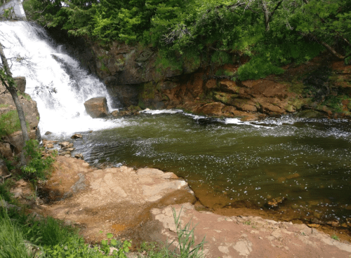 A serene waterfall cascades into a calm pool, surrounded by lush greenery and rocky terrain.