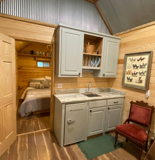 Cozy cabin interior featuring a kitchenette, wooden walls, and a view into a bedroom with a bed and decorative art.