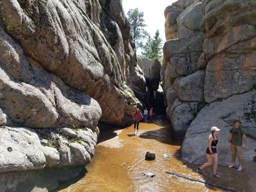 Two people walk through a narrow rocky canyon with a sandy floor, while others explore in the background.