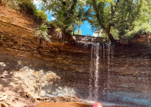 A serene waterfall cascades over rocky cliffs, surrounded by lush greenery and a clear blue sky.