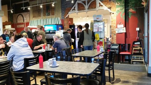 A busy diner scene with patrons at tables, a counter, and a colorful mural in the background. Condiments on tables.