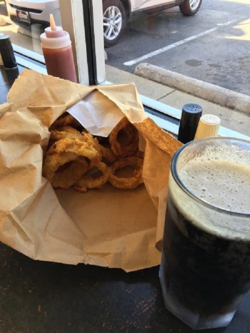 A paper bag with crispy onion rings next to a glass of dark soda on a table by a window.
