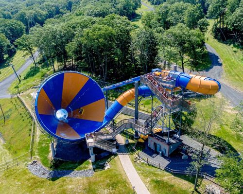 Aerial view of a large water slide with a funnel and twisting tubes, surrounded by trees and grassy areas.