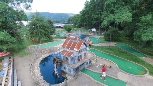 A scenic mini-golf course featuring a blue house, water features, and lush greenery, with players enjoying the game.