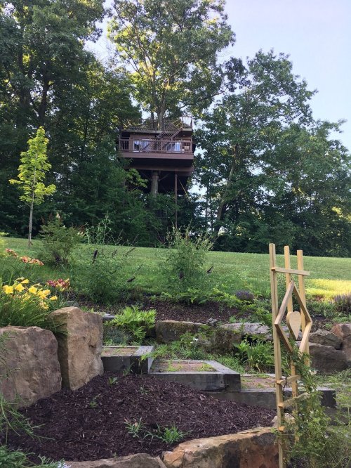A wooden treehouse perched among trees, overlooking a landscaped garden with stone features and colorful flowers.