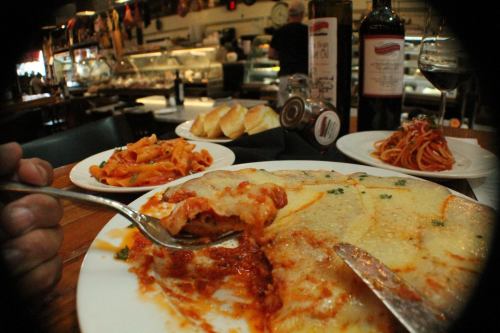 A hand holding a fork with lasagna, surrounded by pasta dishes, bread, wine, and a bustling restaurant background.