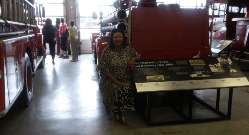 A woman in a patterned dress sits beside a display about the Depression Years in a fire museum, with vintage fire trucks in the background.