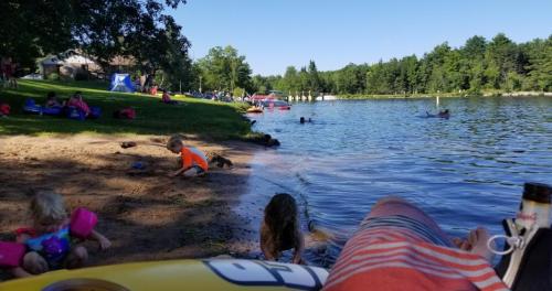 A sunny lakeside scene with children playing on the beach, people swimming, and boats in the water.