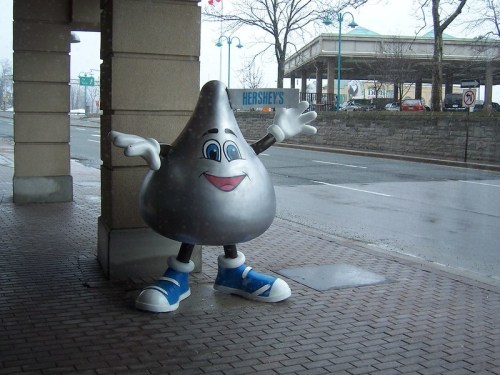 A large, cartoonish Hershey's Kiss character with arms outstretched, wearing blue sneakers, stands by a street.
