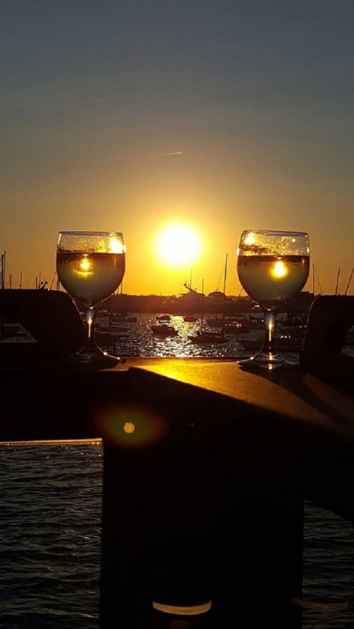 Two glasses of wine sit on a table, reflecting the sunset over a harbor filled with boats.