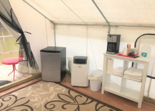 Interior of a tent featuring a small fridge, coffee maker, air conditioner, and a shelf with towels.