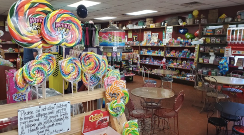 Colorful lollipops in the foreground with a candy shop filled with various sweets and treats in the background.
