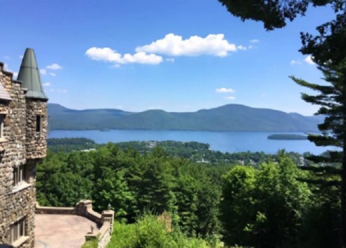 A scenic view of a lake surrounded by mountains, with a stone building in the foreground and blue skies above.
