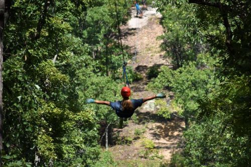 A person zip-lining through a forest, surrounded by green trees, with another person visible in the background.