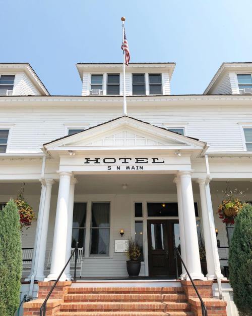 Front view of a charming hotel with a white facade, large porch, and American flag, surrounded by greenery.