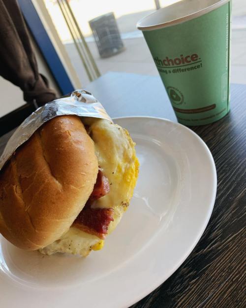 A breakfast sandwich with egg and bacon on a plate, next to a green coffee cup on a table.