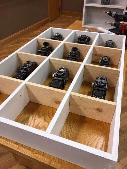 A wooden tray with nine compartments, each holding a vintage camera, placed on a wooden table.