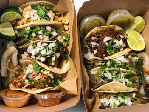 Two takeout boxes filled with tacos topped with onions and cilantro, accompanied by lime wedges and sauces.