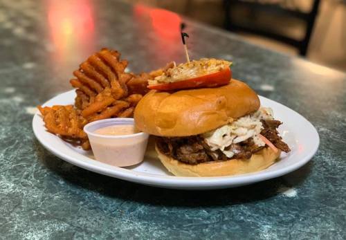 A plate featuring a pulled pork sandwich with coleslaw, a slice of tomato, and sweet potato fries with dipping sauce.