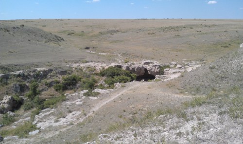 A vast, open landscape with a rocky cave entrance surrounded by sparse vegetation under a clear blue sky.