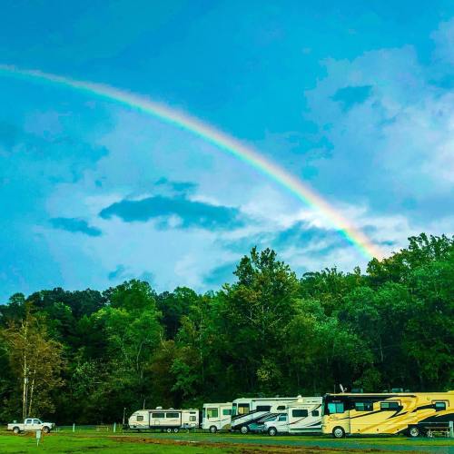 A vibrant rainbow arcs over a row of RVs parked near lush green trees under a partly cloudy sky.