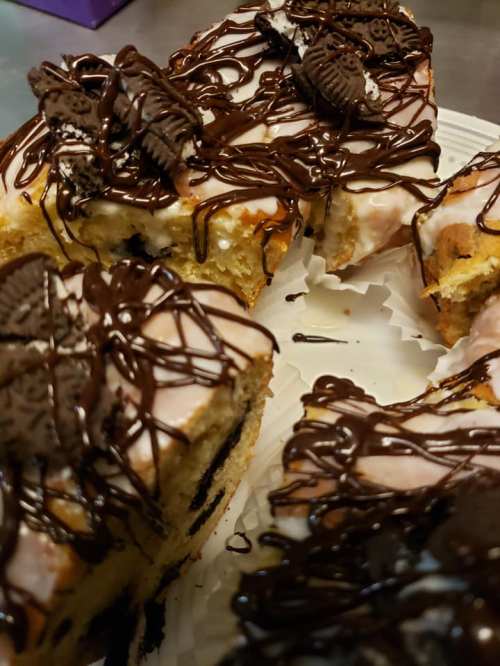 A close-up of a cake topped with chocolate drizzle and Oreo cookie pieces, with a slice removed.