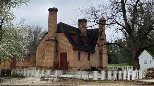 Historic brick house with four chimneys, surrounded by a white picket fence and trees, under a cloudy sky.