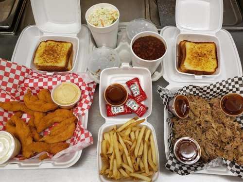 A spread of takeout food including fried shrimp, pulled pork, fries, coleslaw, baked beans, and toast in containers.