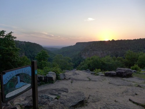Sunset view over a valley from a rocky overlook, with a sign for Cedar Falls Trail in the foreground.