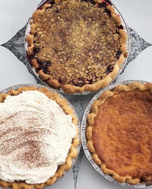 Three pies on a table: a berry crumble, a cream pie topped with whipped cream, and a golden custard pie.