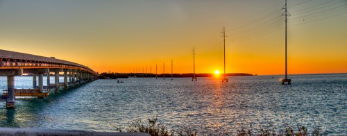 Sunset over a calm body of water, with a bridge and power lines silhouetted against the vibrant orange and yellow sky.