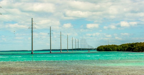 Power lines stretch over turquoise waters, with lush greenery on the shore under a partly cloudy sky.