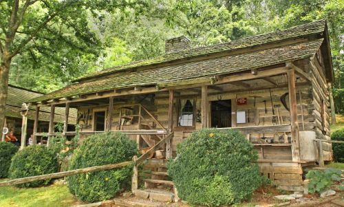 A rustic wooden cabin surrounded by greenery, featuring a porch and various tools displayed outside.
