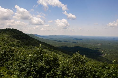 A panoramic view of lush green mountains under a partly cloudy sky, stretching into the distance.