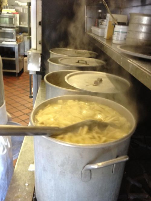Four large pots on a stove, steaming with boiling pasta inside, in a busy kitchen setting.