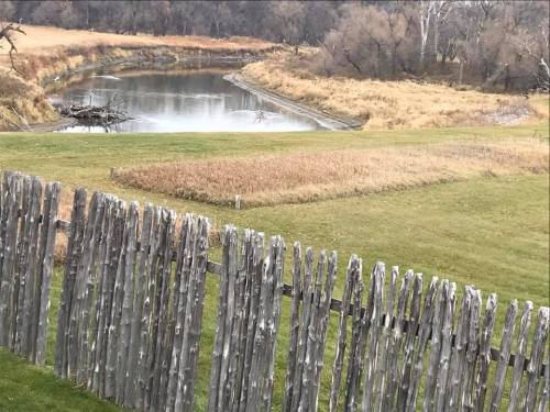 A wooden fence in the foreground with a river and grassy landscape in the background, surrounded by trees.
