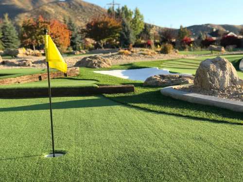 A close-up of a mini-golf hole with a yellow flag, surrounded by green turf and rocky landscaping.