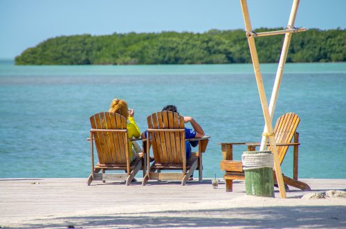 Two people relax in wooden chairs by the water, enjoying the view of a calm sea and distant greenery.