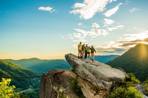 A group of five people stands on a rocky outcrop, overlooking a scenic valley at sunset with vibrant clouds.