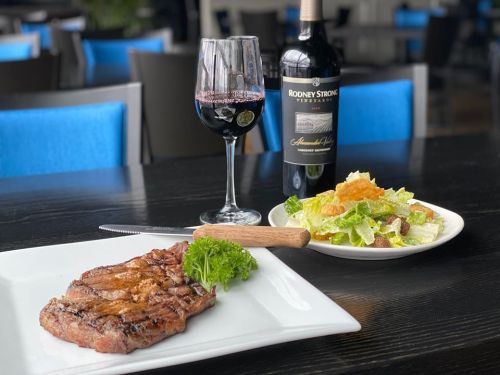 A plate of grilled steak with parsley, a glass of red wine, and a Caesar salad on a table with blue accents.
