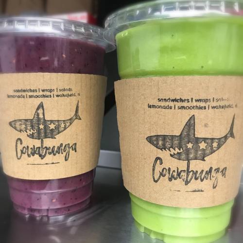 Two smoothie cups with colorful drinks, one purple and one green, both featuring a shark logo and a kraft paper sleeve.