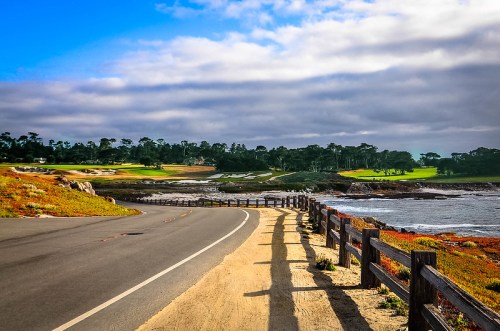 A scenic coastal road curves alongside a golf course, with lush greenery and a cloudy sky in the background.