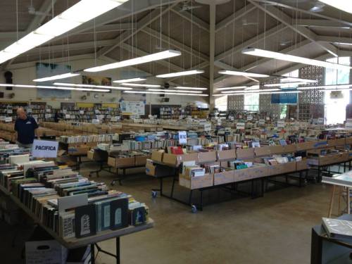 A spacious library filled with rows of tables stacked with books and organized by categories.