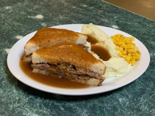 A plate featuring a sandwich with gravy, mashed potatoes, and corn on a green countertop.