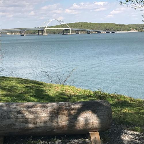 A serene view of a bridge over a calm lake, with green trees in the background and a log in the foreground.